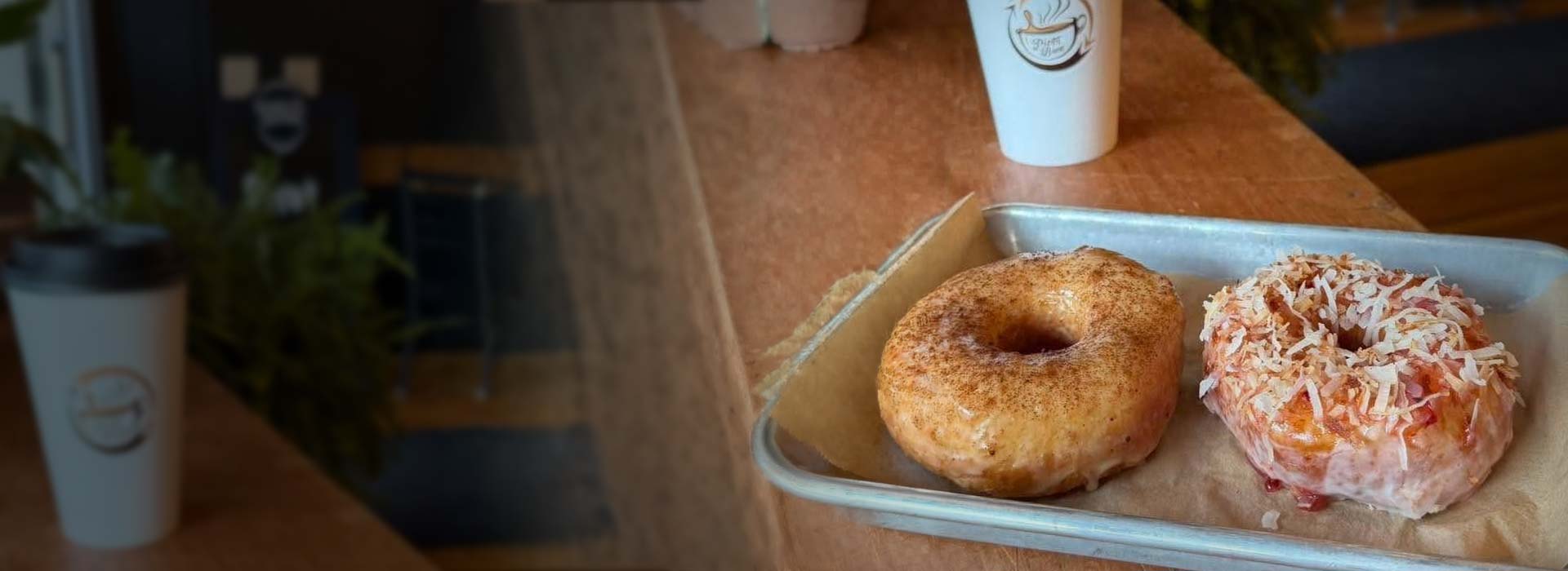 Two glazed donuts on a metal tray next to two coffee cups. Two glazed donuts on a metal tray next to two coffee cups.
